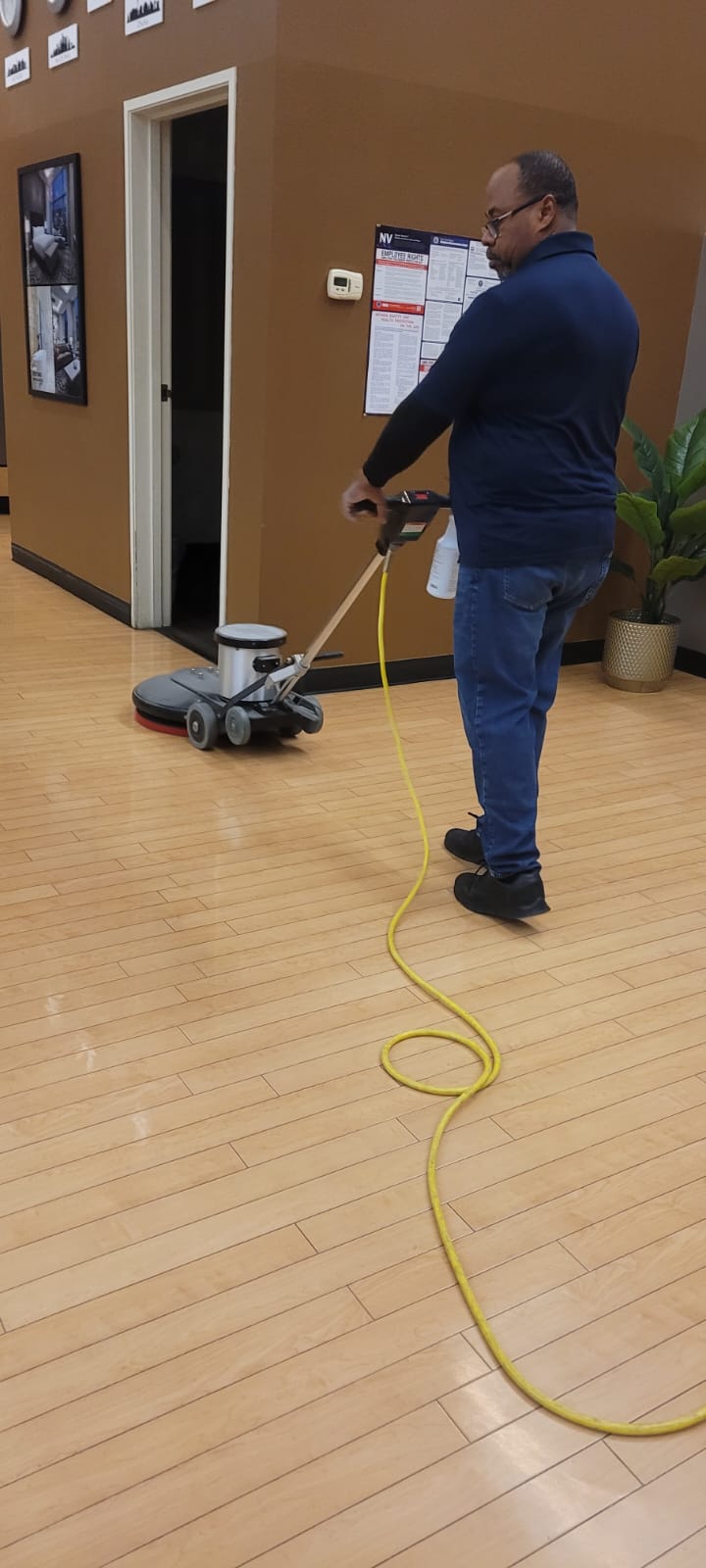 Man using a floor polishing machine on a light wood floor in an indoor space with brown walls.