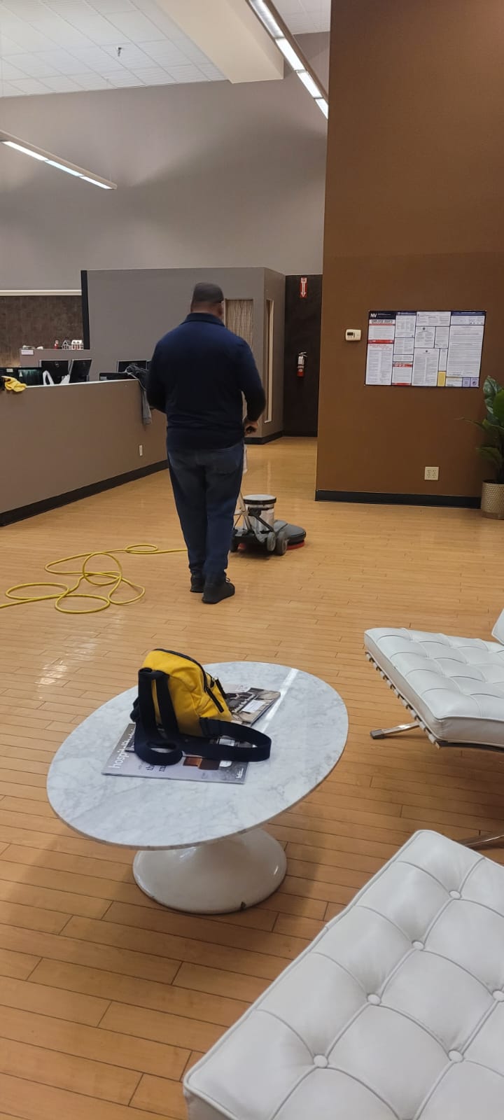 Person cleaning a polished wooden floor with a floor buffer in a modern office with white cushioned chairs and a round marble table with a yellow bag.