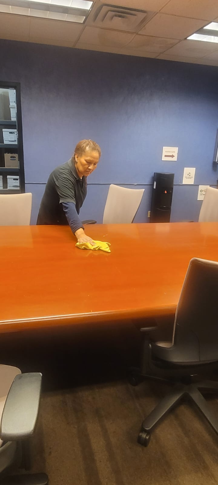 Person cleaning a large wooden conference table with a yellow cloth in an office meeting room.